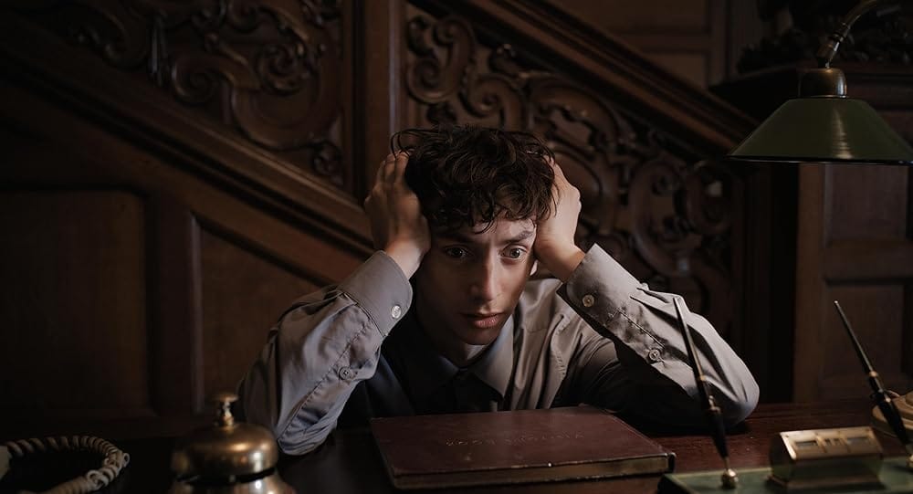 A young concierge leans on the reception desk of a posh hotel with his head of curly brown hair in his hands and a look of shell shock on his face. He is surrounded by bell, ledger and fountainpen trappings and backed by an ornate staircase, but he is lost in his own thoughts.