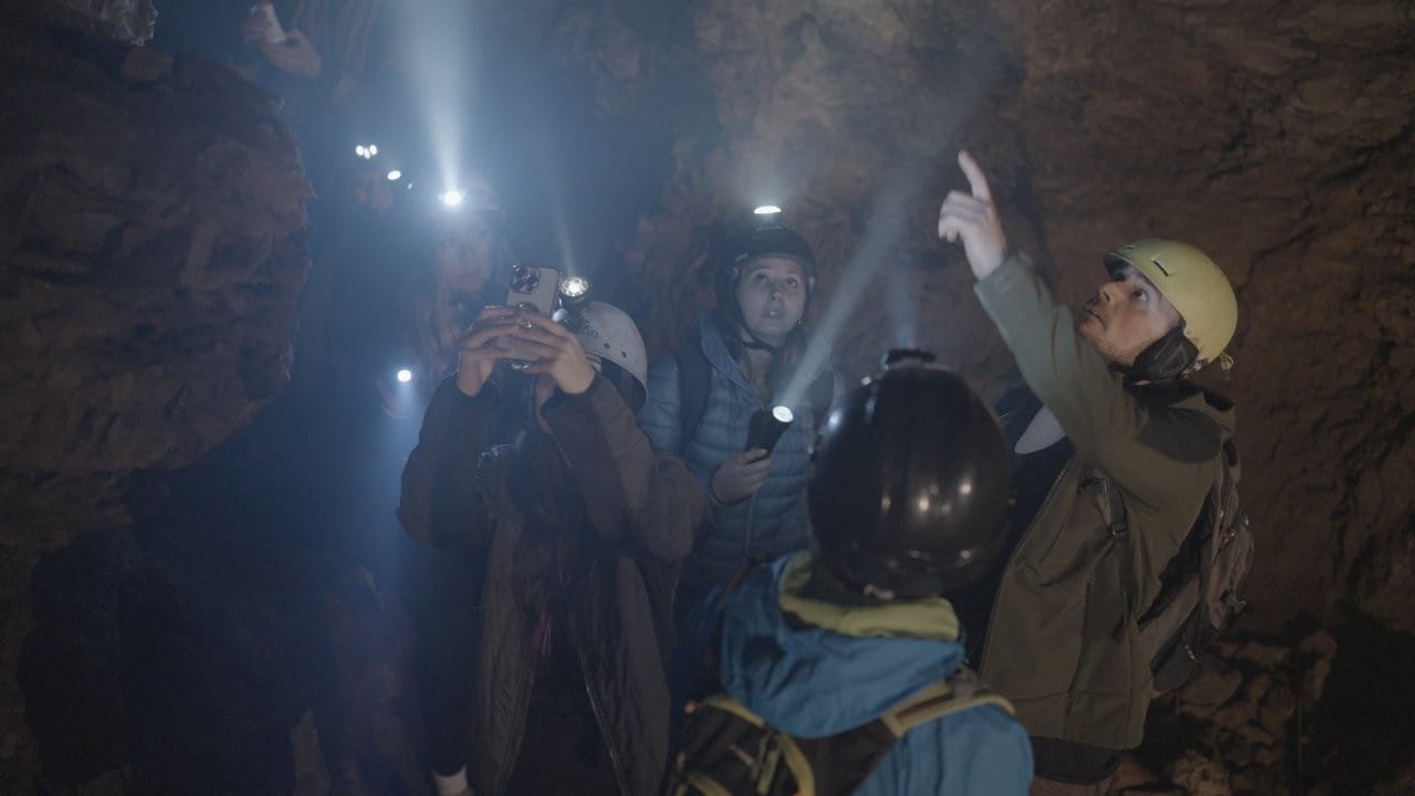 A large crowd of men and women in caving gear stand in a crevass-filled cave, lights shining from their hands and helmets. One guy points up and another lady tries to video the ceiling on her phone. Olivia (centre) looks dubious.