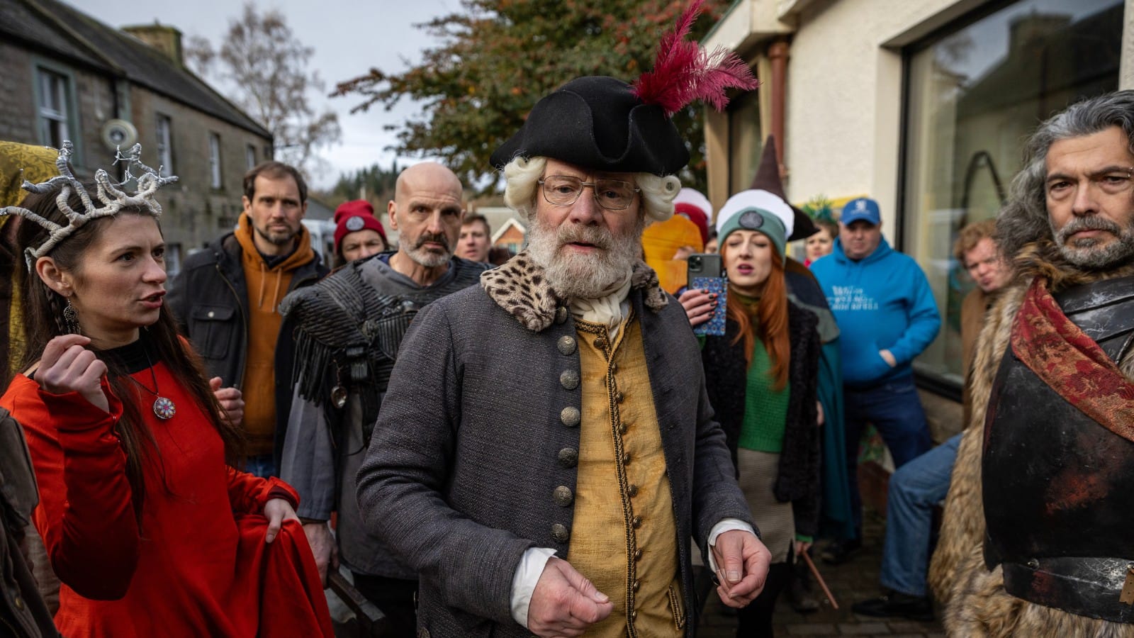 An elder man dressed in Scottish enlightenment era clothing stands concerned and bemused amidst a line of fans cosplaying for their favourite high fantasy show. They look displeased at his prescence.